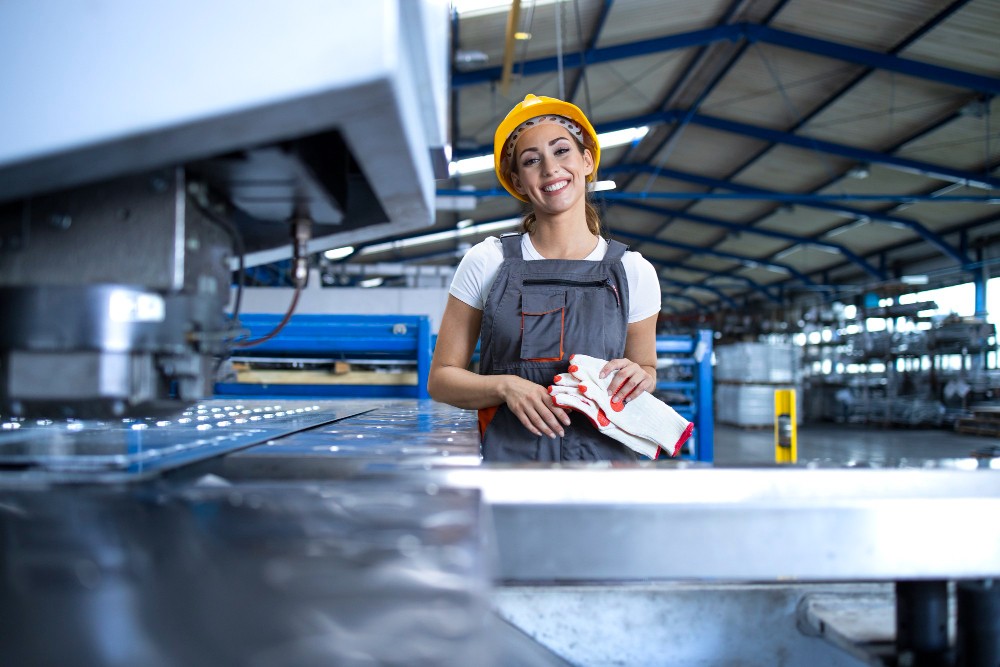 Operaia Di Fabbrica Con Indumenti Protettivi E Casco In Piedi Accanto A Una Macchina Industriale Nella Linea Di Produzione